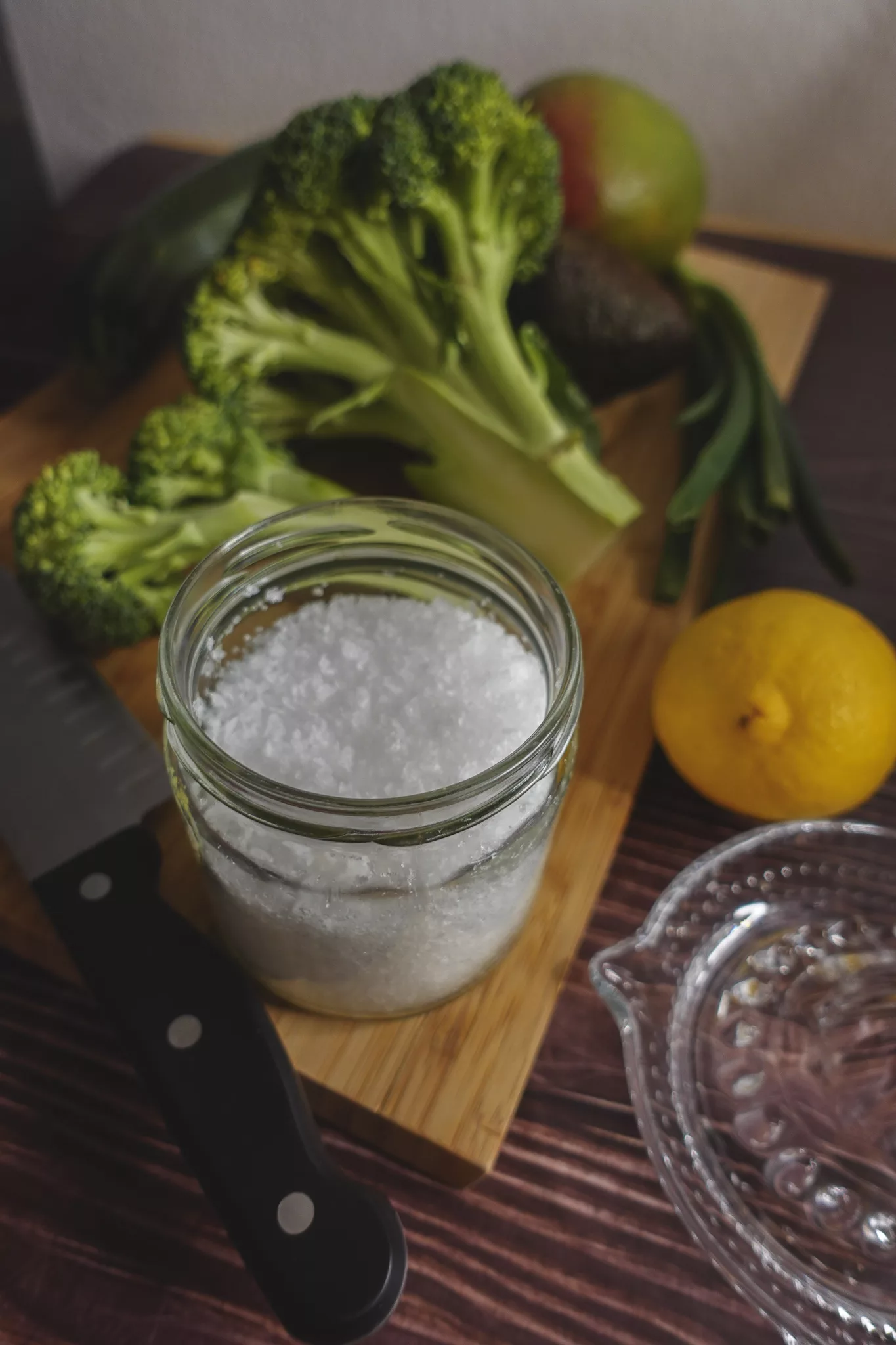 Salz, Zitrone und Brokkoli auf einem Holzschneidebrett. Links vorne liegt ein Messer, rechts vorne eine Zitronenpresse aus Glas.